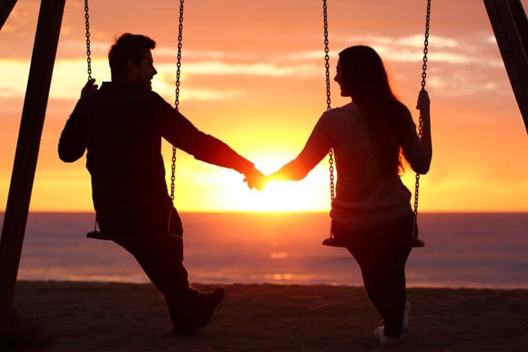 Back light portrait of a couple silhouette sitting on swing holding hands watching a sunrise on the beach with the sun in a warmth background