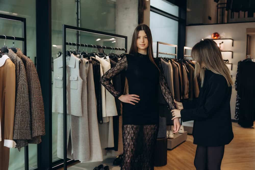 Female client standing in front of clothing rack, personal stylist adjusting sleeve of her elegant black lace dress in boutique