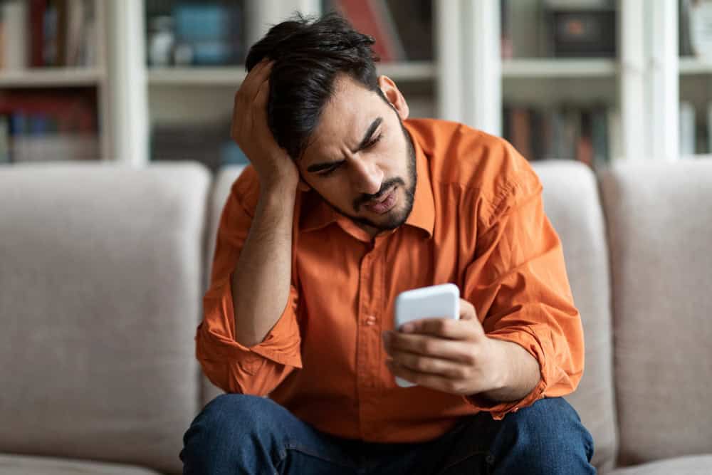Anxious upset shocked arab guy in casual sitting on couch at home, holding smartphone, looking at ga