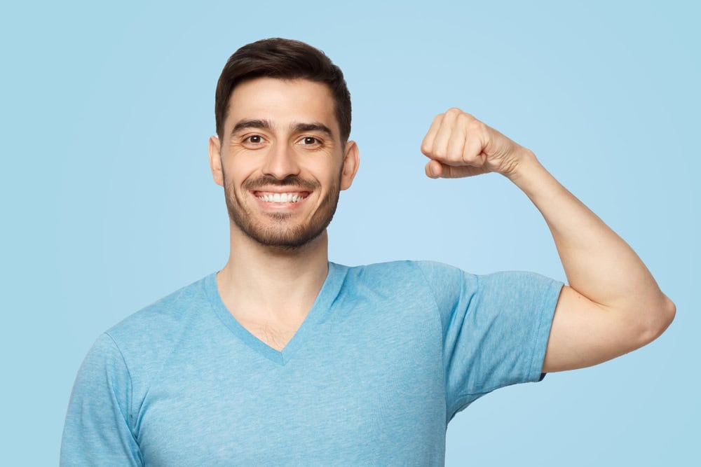 Young strong sporty athletic man in casual t-shirt, showing biceps after training in gym, isolated on blue background