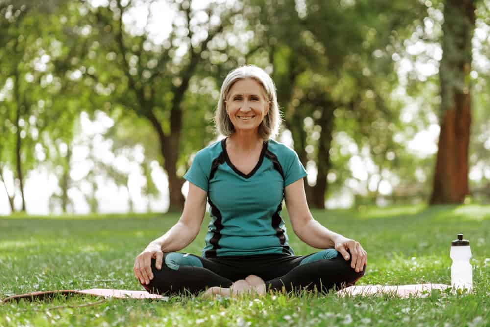 Smiling beautiful senior woman practicing yoga in park sitting on yoga mat, outdoors. Balance, harmony concept