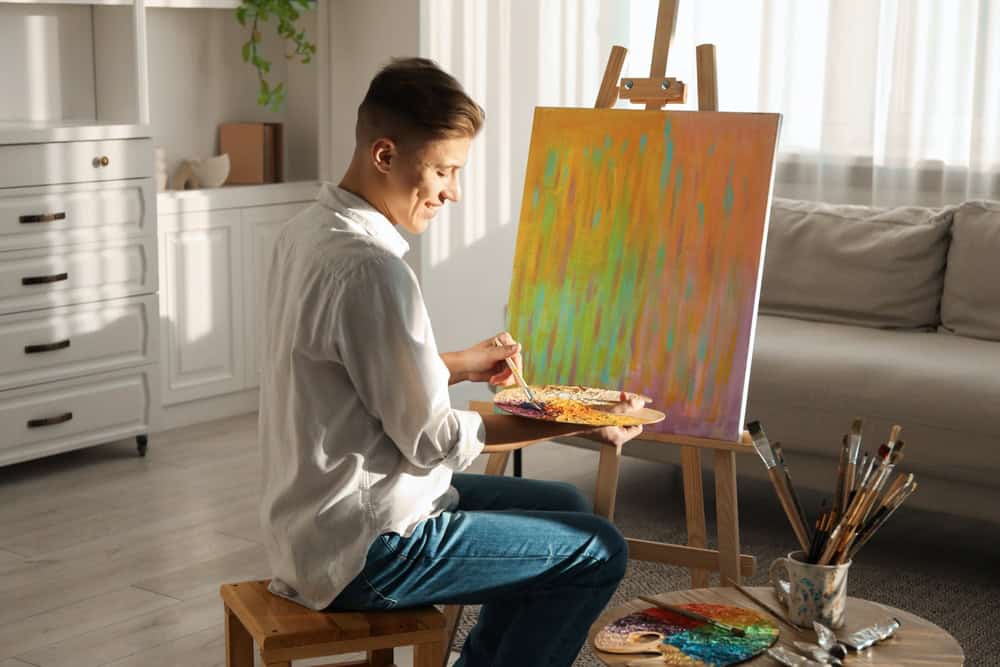 Smiling man with palette and paintbrush drawing abstract picture at home