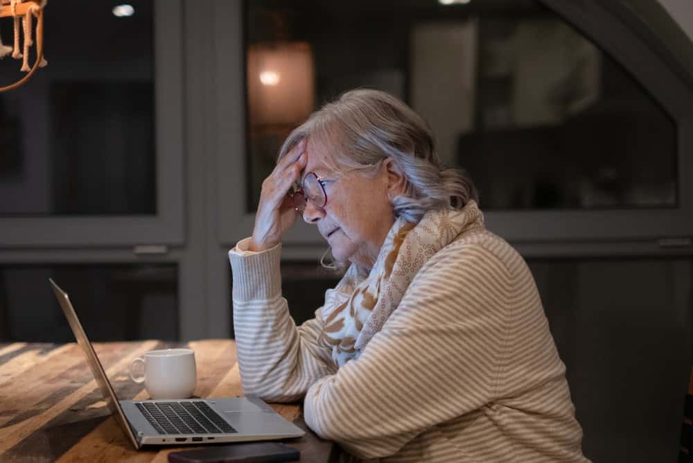 Senior woman using a laptop at home in the evening, sitting at a wooden table under warm light, representing digital literacy, online communication, and independent lifestyle for older adults