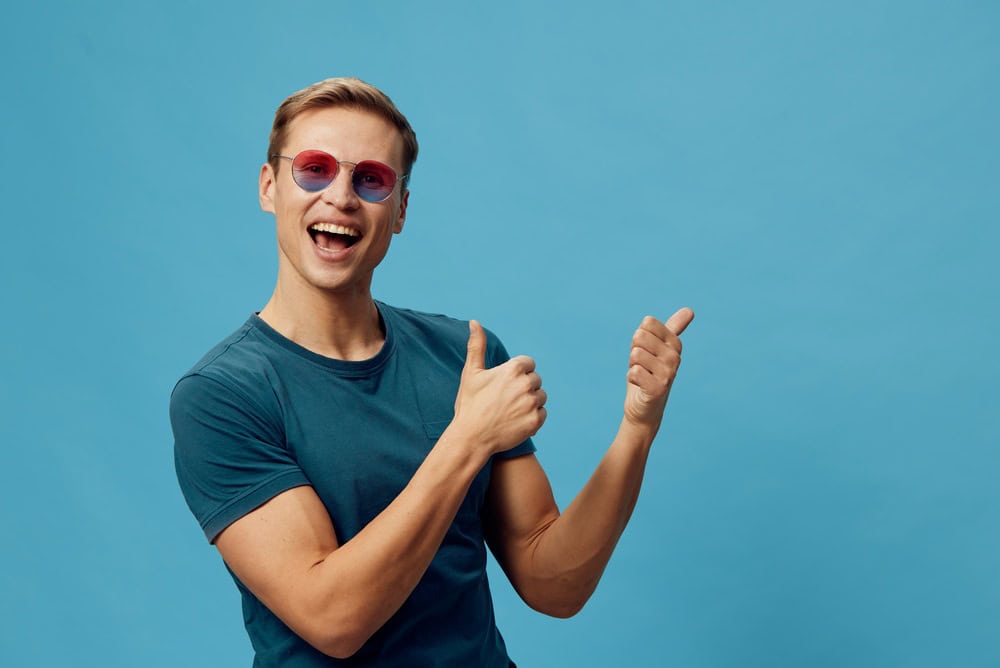 Cheerful young man wearing sunglasses and a blue T-shirt showing thumbs up gesture against a plain blue background. Happy, casual, positive vibe, studio portrait, lifestyle concept, summer fashion