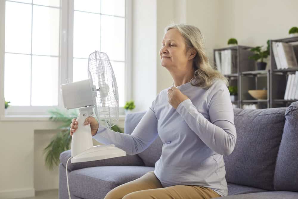 Senior older woman sitting on sofa, holding and directing fan, fanning her face, appearing flushed and uncomfortable, experiencing heat flash, indoor living room setting, bad hot room climate