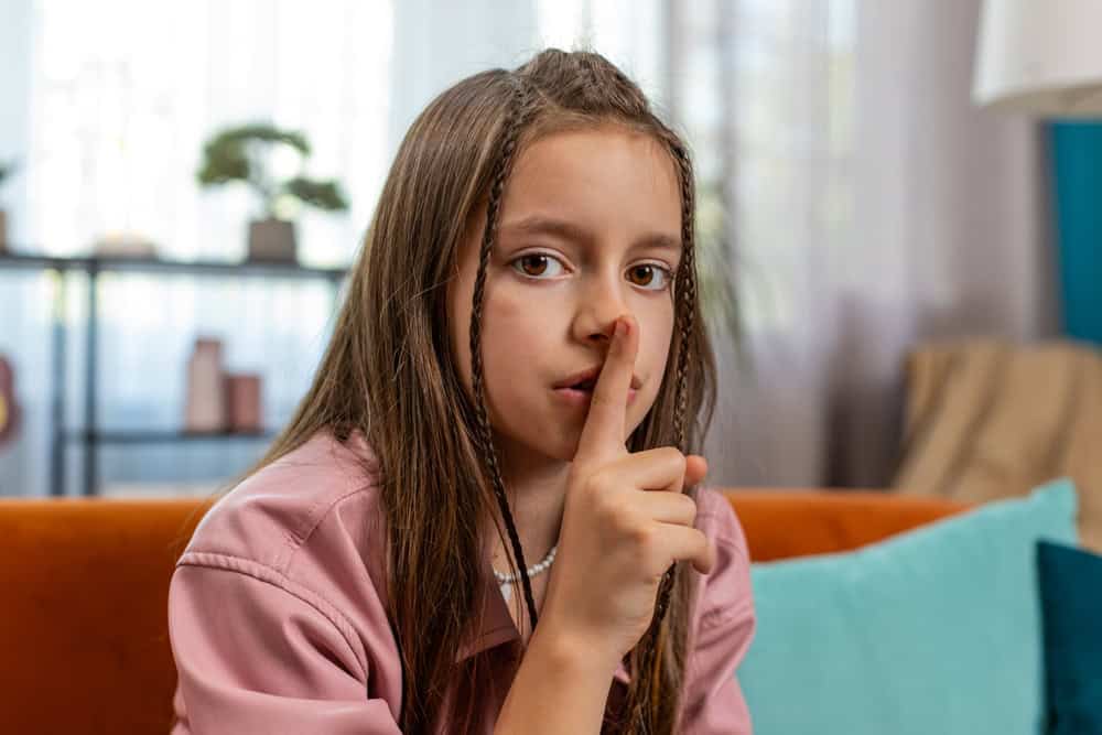 Shh be quiet please. Portrait of preteen girl child presses index finger to lips makes silence hush gesture sign do not tells gossip secret. Little cute teenage female sitting on sofa in living room.