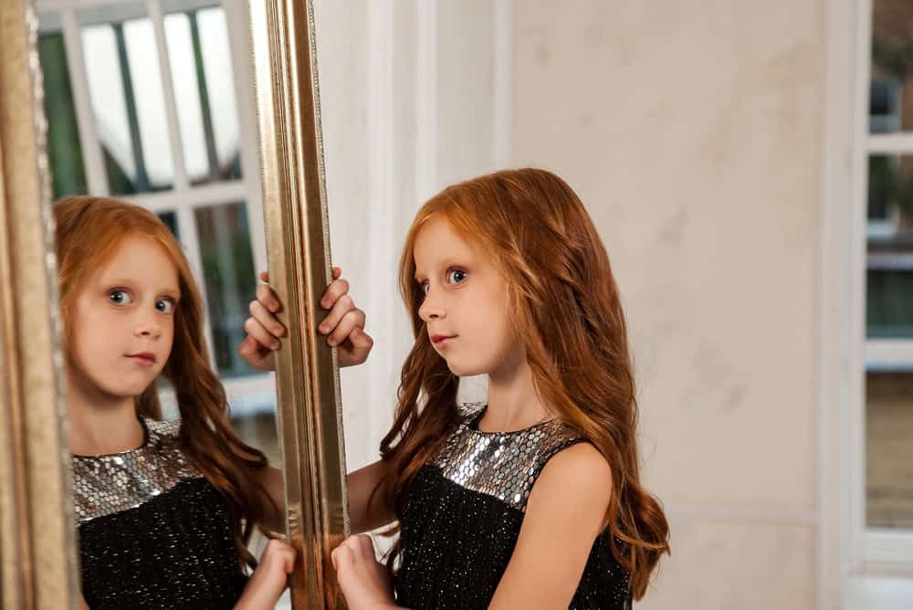 A vibrant red-haired girl, adorned in a sparkling black dress, gazes thoughtfully into an ornate golden mirror.