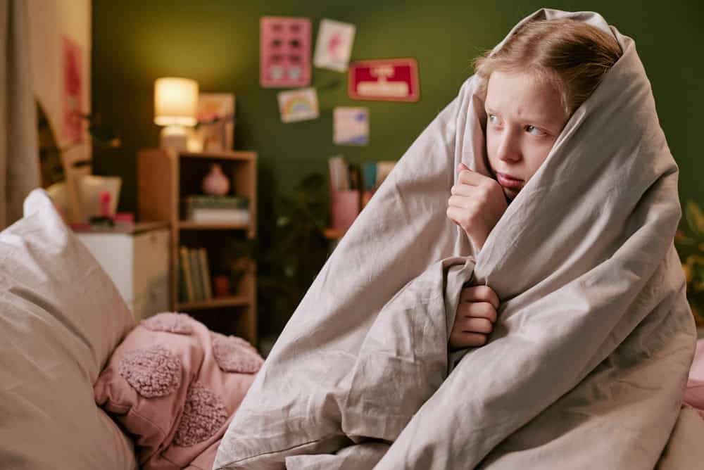 Caucasian child girl sitting on bed wrapped in blanket looking to side with worried expression, surrounded by pillows and bedroom decor, showing concern or fear in nighttime setting