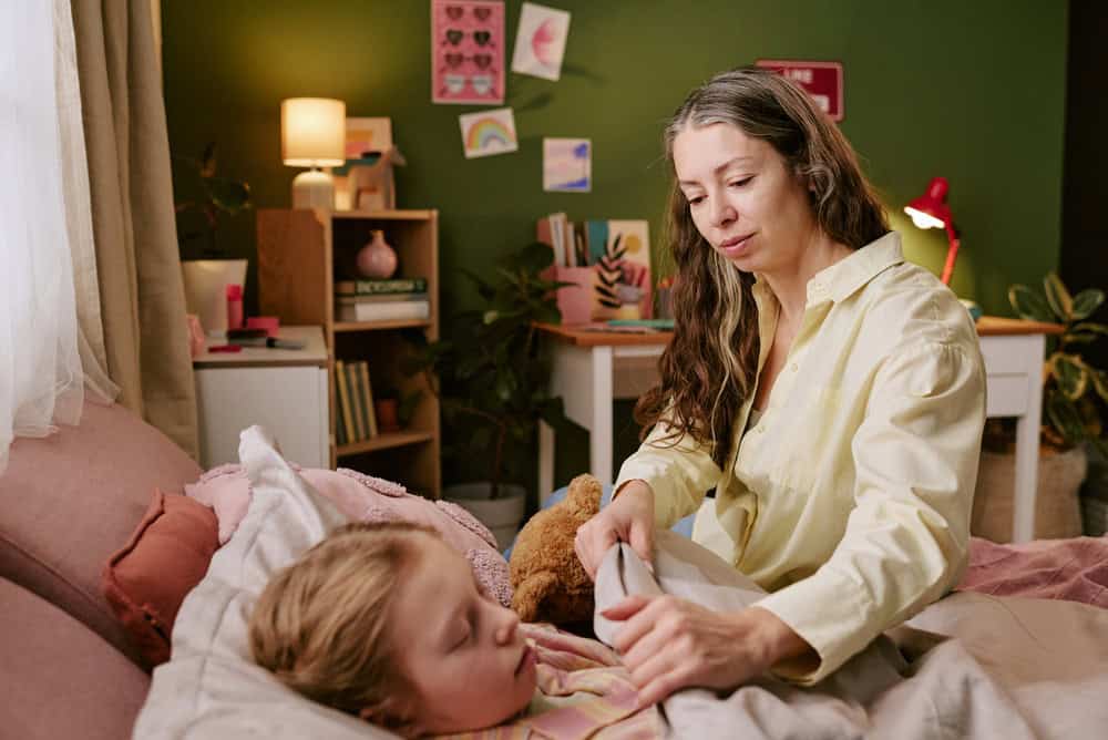 Middle aged Caucasian woman tucking in sleeping Caucasian child in bed, gently arranging blanket while sitting beside bed in cozy bedroom decorated with children's drawings and toys