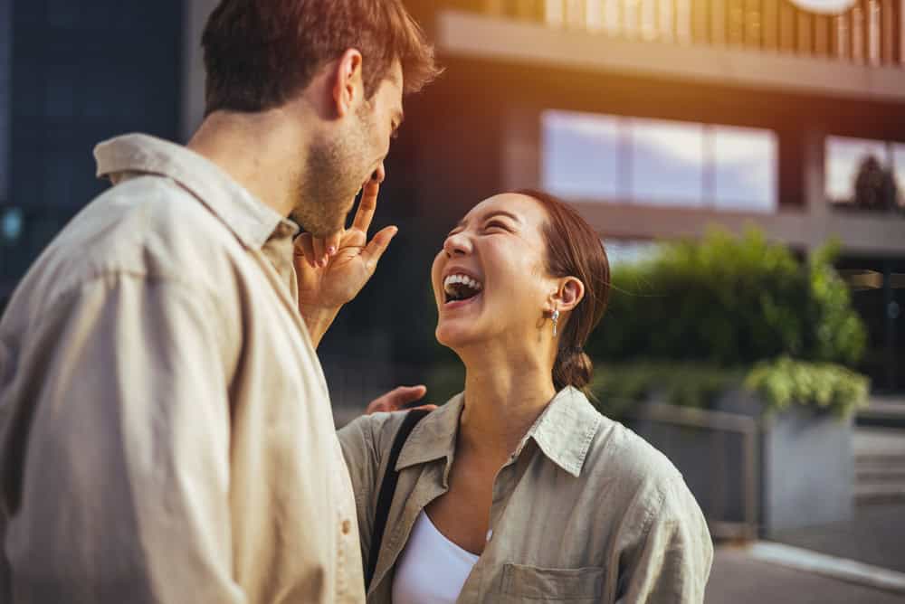 A young couple share a joyful, playful moment laughing together outdoors in an urban plaza, conveying affection, connection and lighthearted romance on a sunny day
