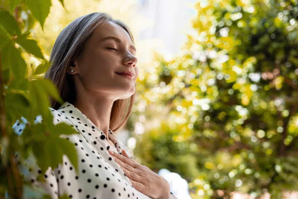 Young office worker taking a deep breath with closed eyes, enjoying a moment of mindfulness in a green park, connecting with nature