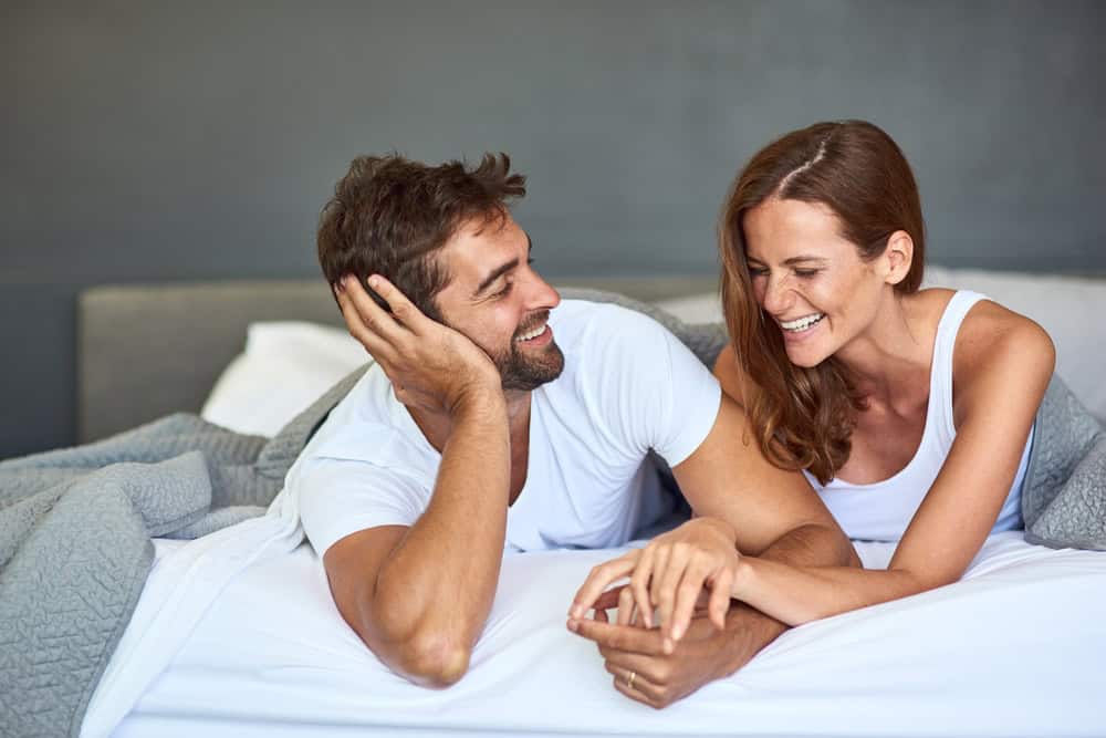 Happy couple, wake up and laughing in bedroom, morning or support for marriage commitment at house. Love, bonding or funny people with smile for romantic relationship, joke or anniversary together