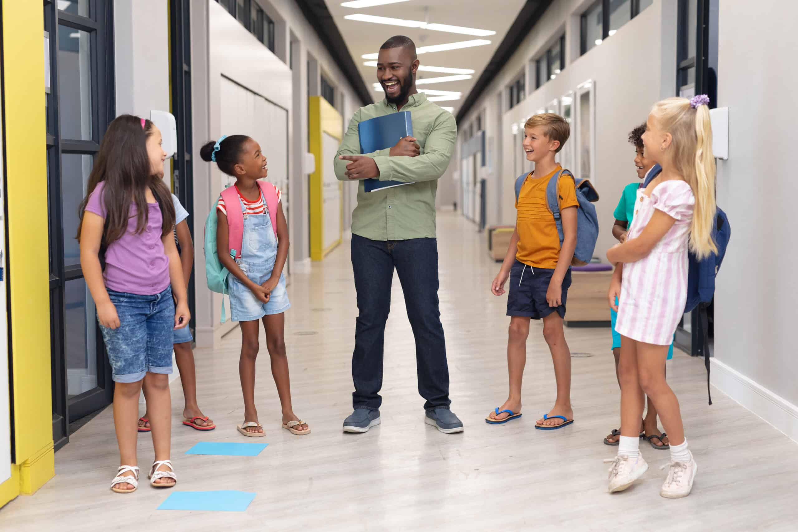 African american young male teacher talking to multiracial elementary students standing in corridor. unaltered, education, learning, teaching, communication, occupation and school concept.
