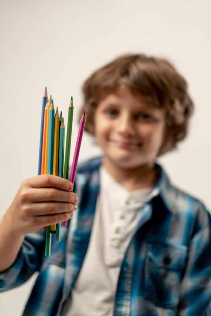 Close up in photo studio on a white background stands a smiling schoolboy in a white sweater and a blue checkered shirt with colored pencils love for drawing