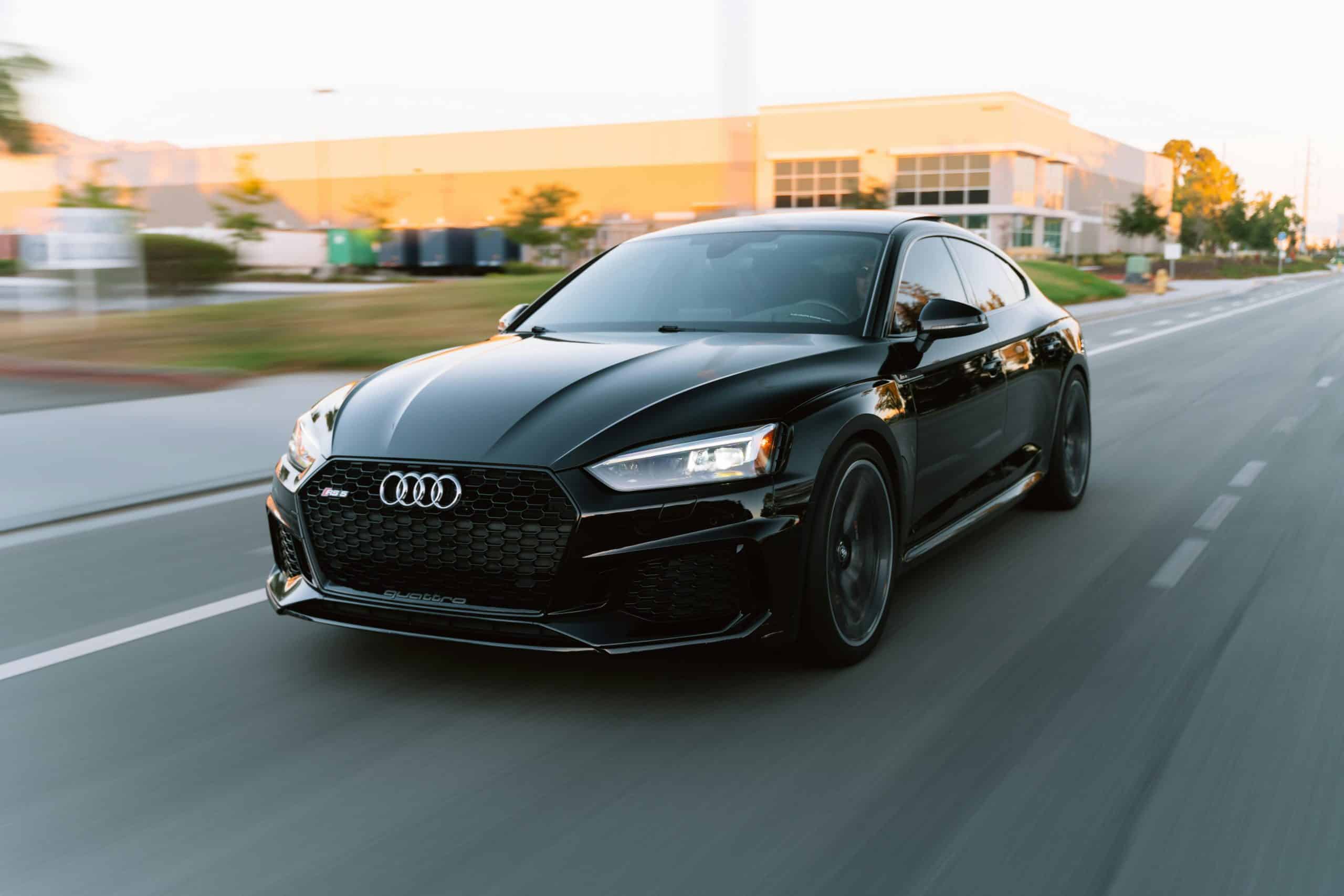 Black Audi sports car driving on city street at sunset.