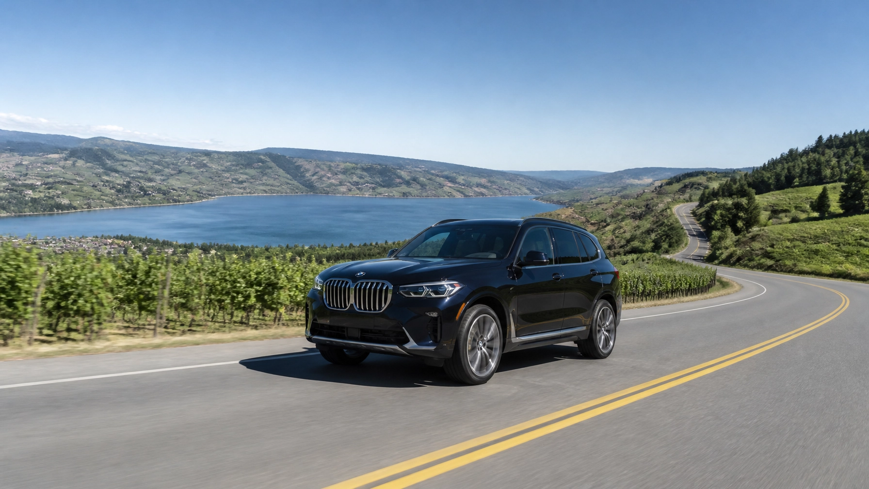 Black SUV driving on a winding mountain road with scenic lake and hills in the background.