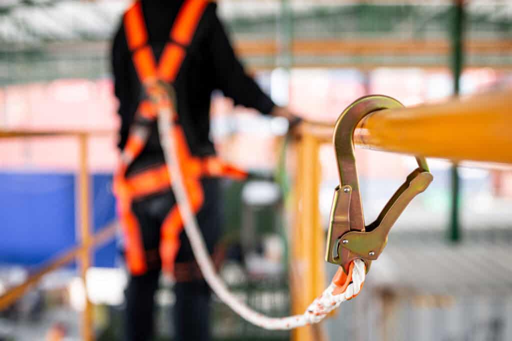 A close-up image of a safety harness and carabiner secured to a yellow safety railing, with a worker wearing an orange harness blurred in the background. The harness and carabiner symbolize the importance of fall protection and safety measures in the workplace. The worker, although not in focus, is seen standing on a platform, indicating an elevated work environment. This image emphasizes the necessity of safety equipment and procedures in preventing accidents, particularly in industries where working at height is common.
