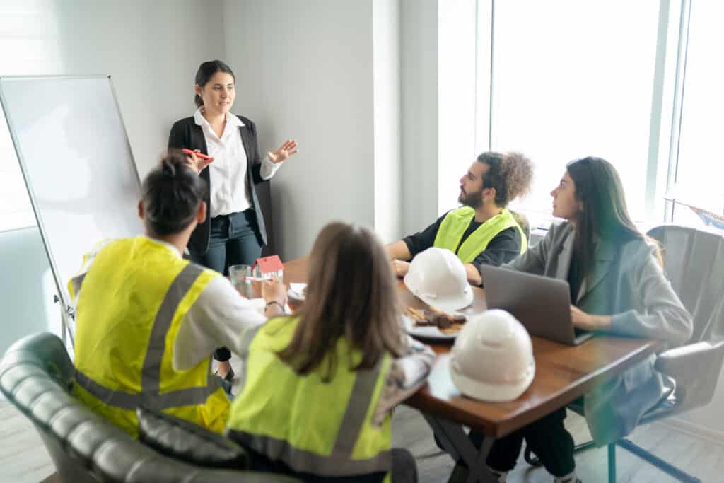 Construction safety training session in progress, featuring a female instructor presenting to a group of construction workers wearing high-visibility vests and hard hats. A whiteboard and table with safety equipment are visible, emphasizing practical, hands-on learning.