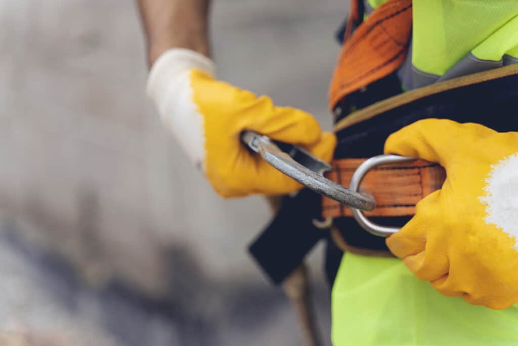 Worker fastening a safety harness with protective gloves on a construction site, symbolising compliance with UK health and safety regulations such as the Work at Height Regulations 2005.