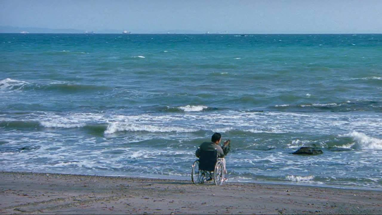 Ren Osugi in HANA-BI sitting in a wheelchair watching the ocean, holding seaweed