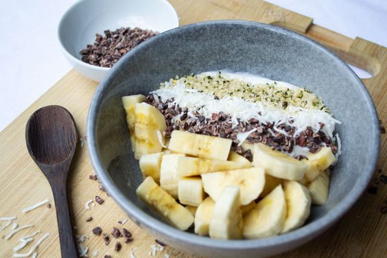 Rich banana and shredded coconut smoothie bowl topped with cacao nibs, served in a rustic gray bowl with a wooden spoon on a wooden cutting board.
