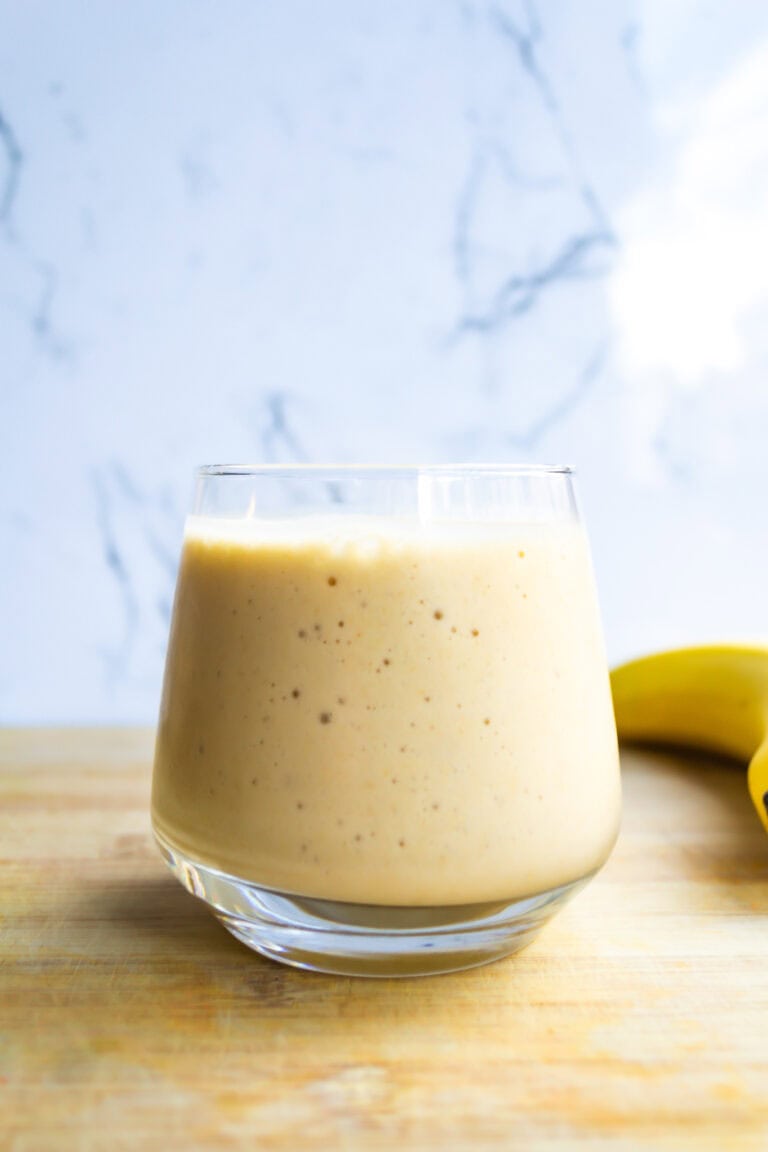 Creamy banana smoothie with chia seeds in a clear glass on a wooden surface with a blue textured background.