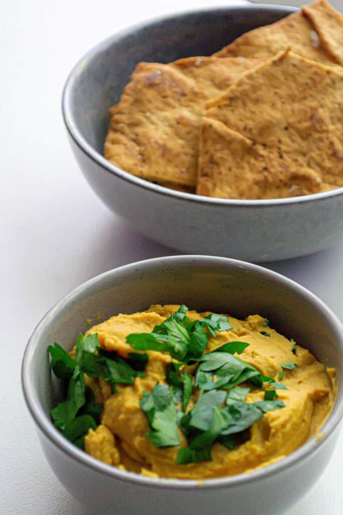 Creamy hummus garnished with fresh parsley and served with crispy pita chips in a gray bowl. Perfect for healthy snacking and appetizer platters.