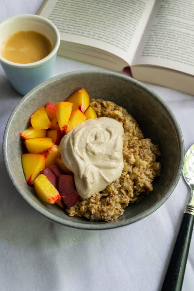 Creamy oatmeal topped with whipped cinnamon almond butter, served alongside fresh peach and cherry plum slices, with a cup of coffee and an open book for a wholesome breakfast experience.
