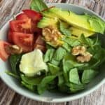 Fresh green spinach, ripe tomatoes, creamy mozzarella, sliced avocado, walnuts, and balsamic dressing in a white ceramic bowl on rustic wooden table for healthy vegetarian salad.