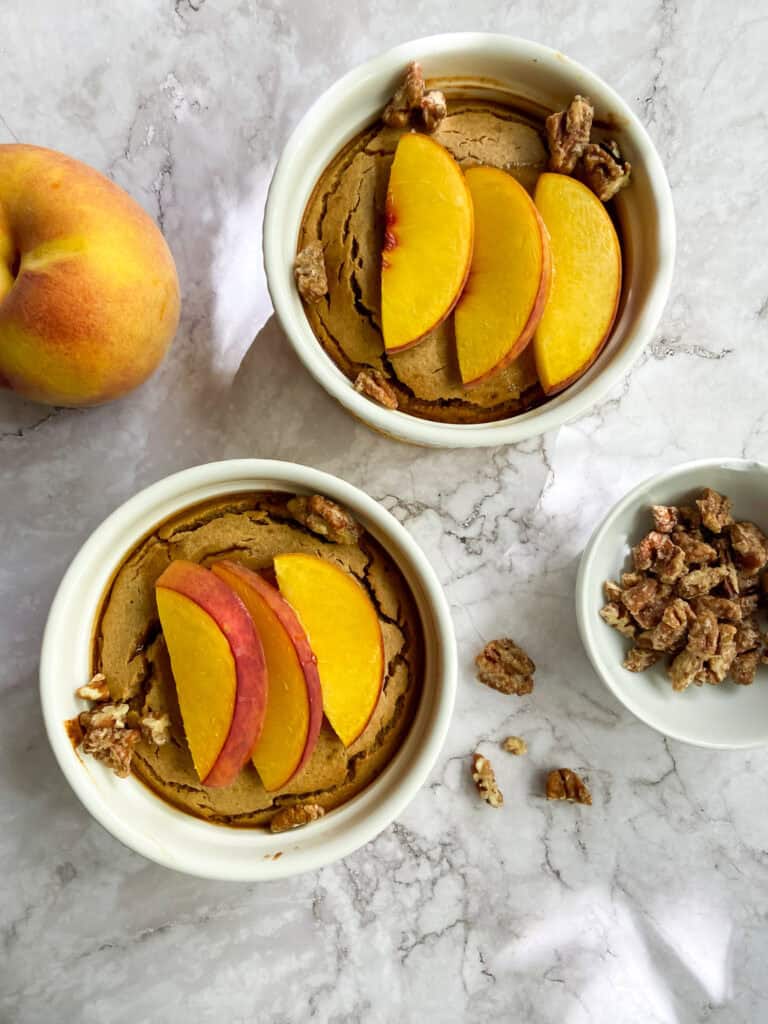 Sweet Potato Pie topped with fresh peach slices and chopped walnuts, served in white ramekins on a marble surface. A whole peach and extra walnuts are also included for added flavor and presentation.