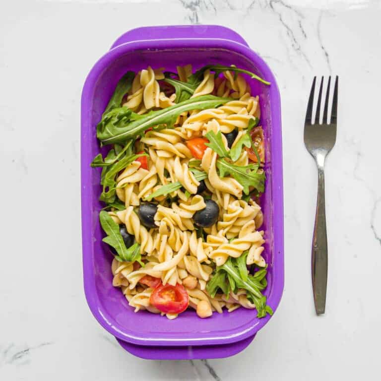 Creamy vegan pasta salad with fresh cherry tomatoes, black olives, and arugula in a purple container on a white marble surface. Perfect for healthy, quick lunch ideas.