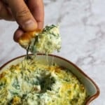Cheesy spinach and artichoke dip being served with a bread piece, cheesy and creamy texture, fresh ingredients, perfect for appetizer or snack, popular party dish, addictive cheesy dip, simple fresh ingredients, flavorful and satisfying.