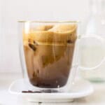 Creamy iced coffee with frothy top in a clear glass mug, served on a white saucer with coffee beans, fresh milk in a small jar, perfect for coffee lovers.