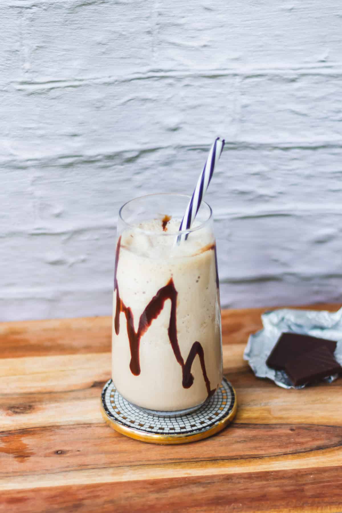 Sweet vanilla milkshake with chocolate syrup in a tall glass on wooden table near water body, striped straw, cold summer drink, refreshing dessert, delicious milkshake for warm weather, Simply Fresh Foodie.