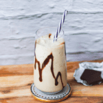Creamy banana smoothie with chocolate drizzle in a tall glass on a wooden table near water.