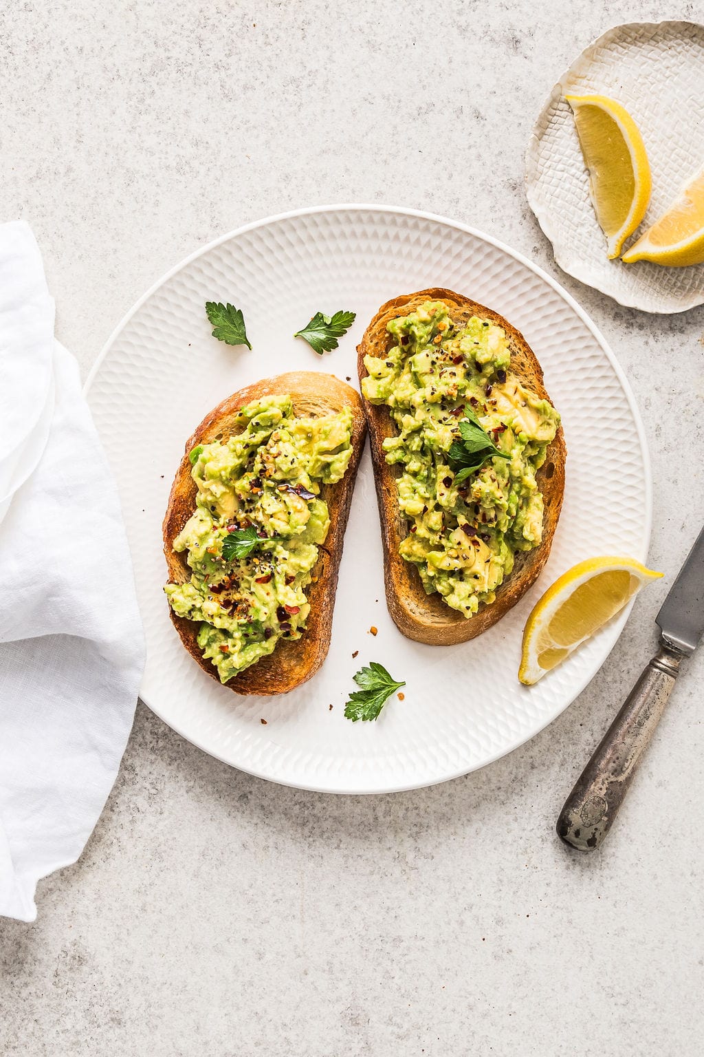 Creamy avocado toast with lemon wedges on a white plate, garnished with fresh herbs and crushed red pepper flakes, perfect for a healthy breakfast or snack.