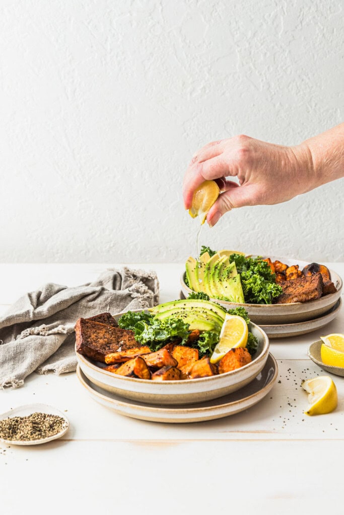 Front view of a hand squeezing lemon juice over the top of a sweet potato tofu nourish bowl.