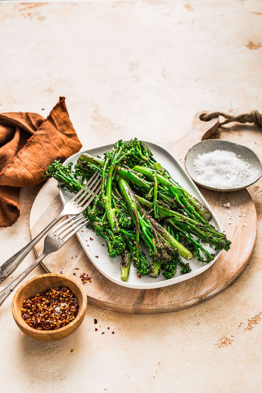 Steamed broccolini seasoned with red pepper flakes and sea salt on a white oval plate, garnished with sesame seeds, served on a round wooden board with salt, chili flakes, and rustic napkins for healthy eating.