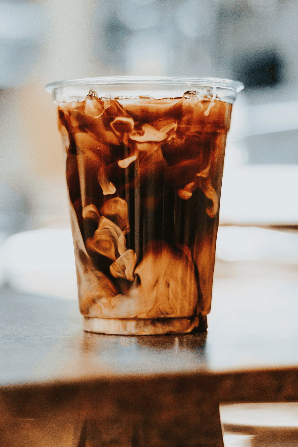 Creamy iced coffee with swirling milk, served in a clear plastic cup on a wooden table. Refreshing summer drink showcasing coffee, milk, and ice.