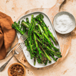 Broiled broccolini with red pepper flakes, sea salt, and garlic on a white platter on a wooden cutting board with seasonings and cloth napkin.