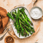 Grilled broccolini on a white platter with red pepper flakes, salt, and utensils, served on a rustic wooden cutting board; healthy vegetable side dish with fresh, vibrant greens.