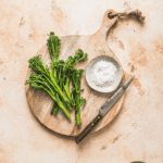 Fresh broccolini on a wooden cutting board with sea salt and a knife, ideal for healthy cooking recipes and nutritious meal prep.