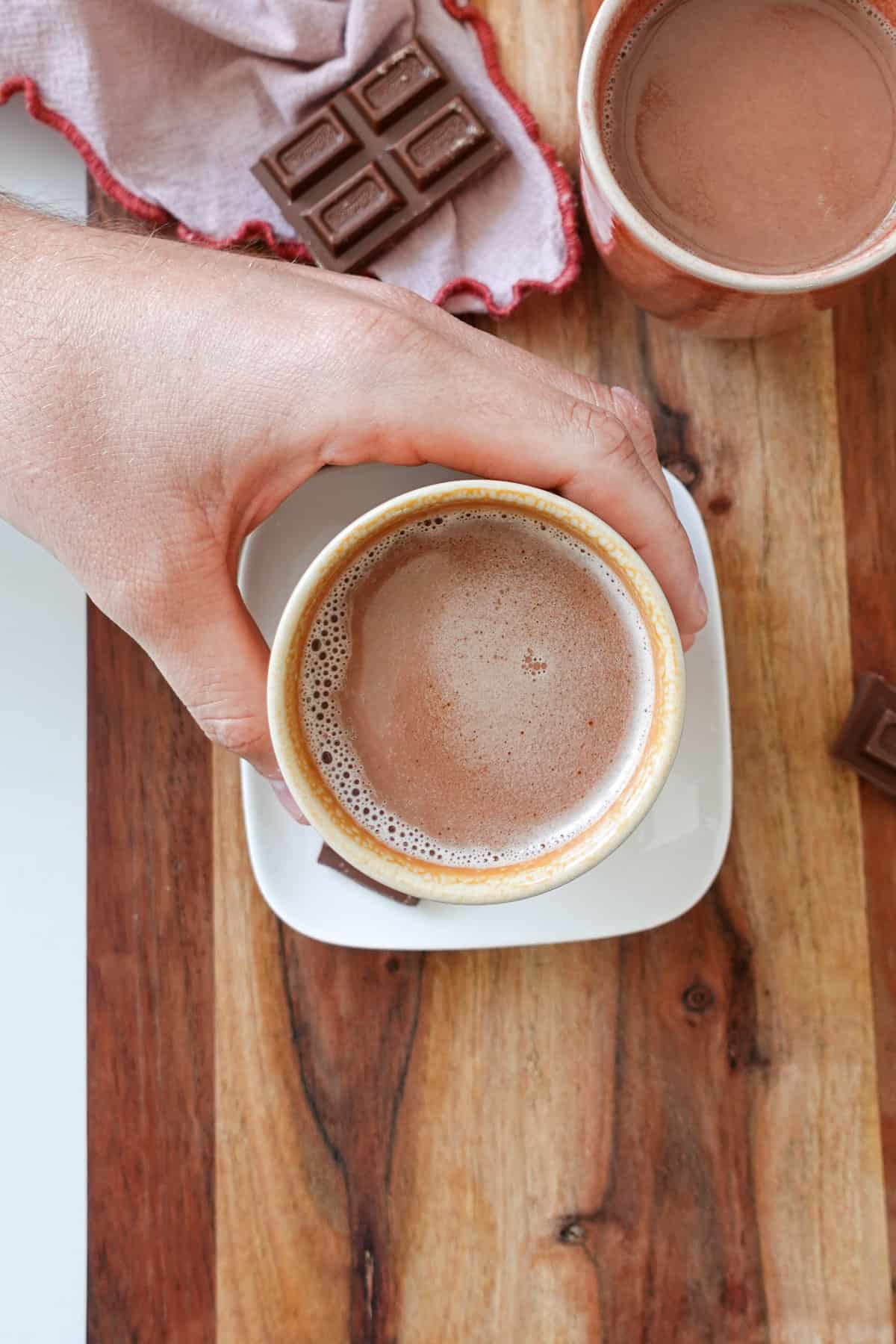 An overhead shot of a hand reaching in and grabbing a mug of Biscoff hot chocolate with another mug and chocolate pieces in the background.