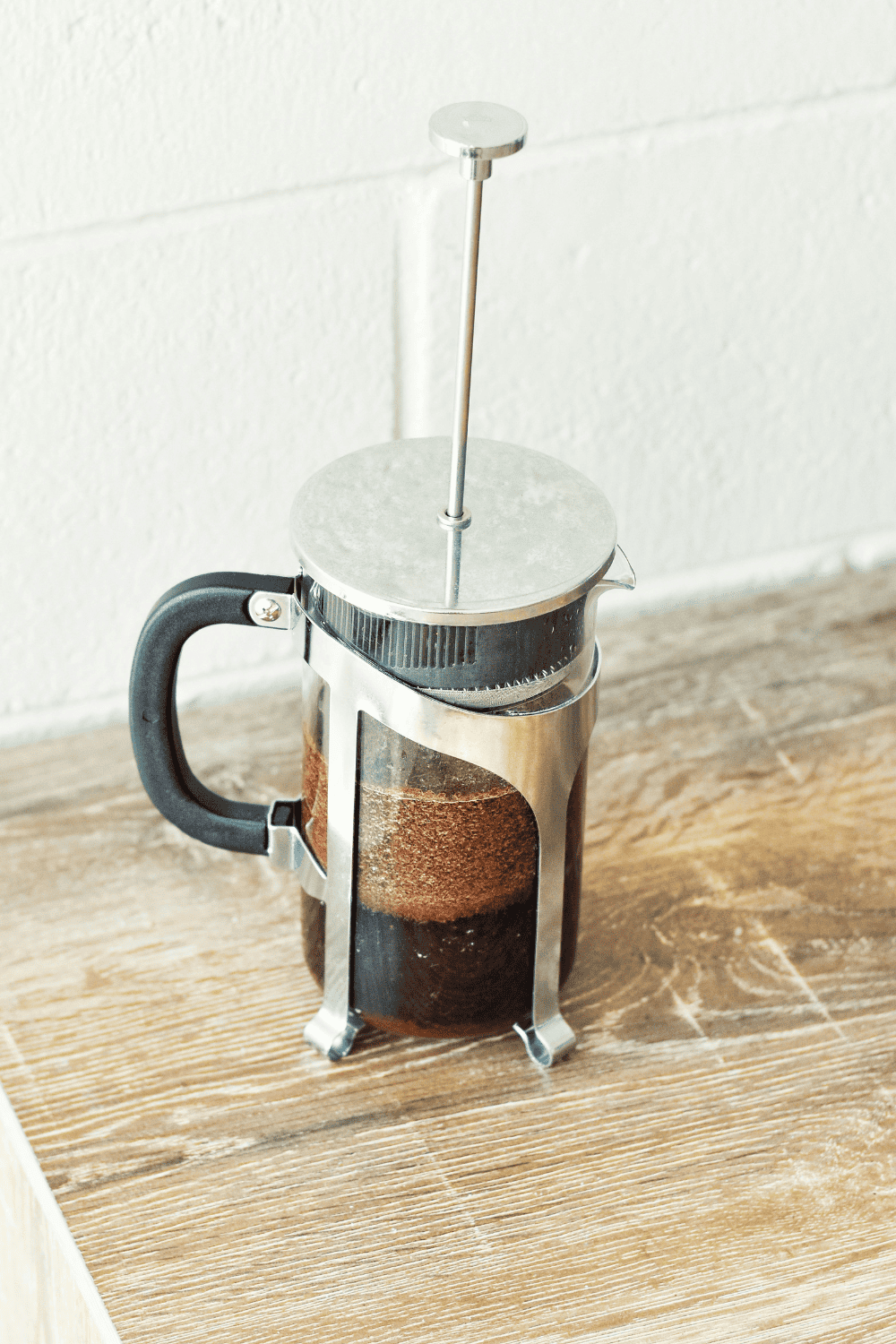 Freshly brewed French press coffee with coffee grounds visible through the glass carafe, placed on a rustic wooden surface in a cozy kitchen setting.