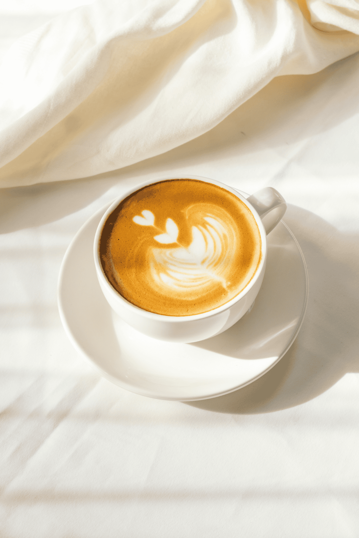 Crema coffee with latte art, served in a white cup on a matching saucer, featuring a heart and floral design, on a white tablecloth with natural lighting.