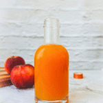 Homemade peach syrup in a glass bottle with fresh peaches in the background for delicious summer beverages and dessert toppings.