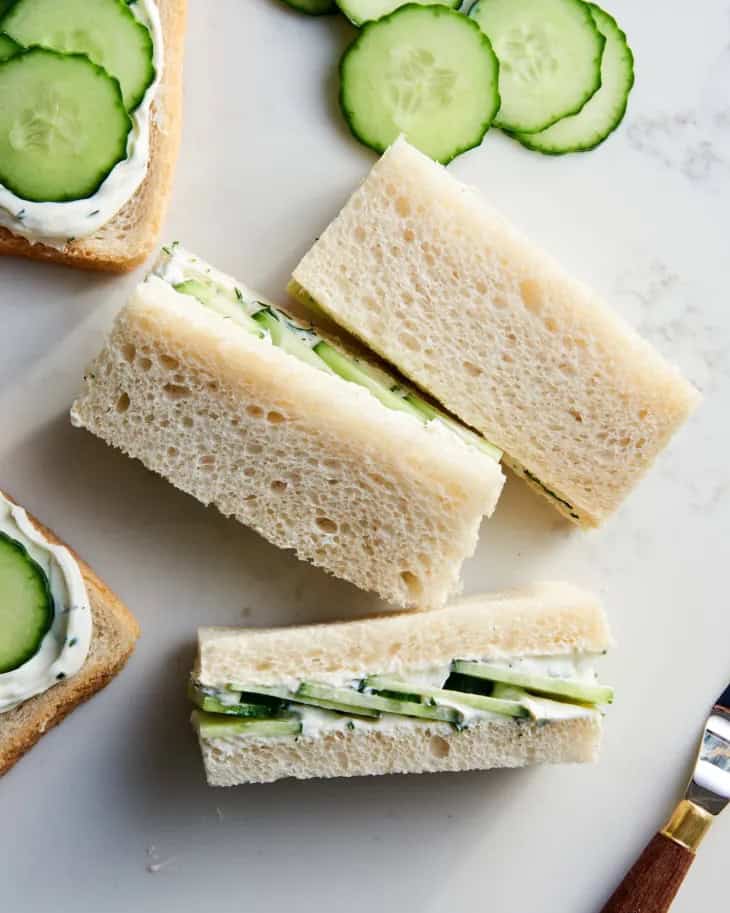 Fresh cucumber sandwiches on white bread with cream cheese, on a white marble surface, garnished with sliced cucumbers.