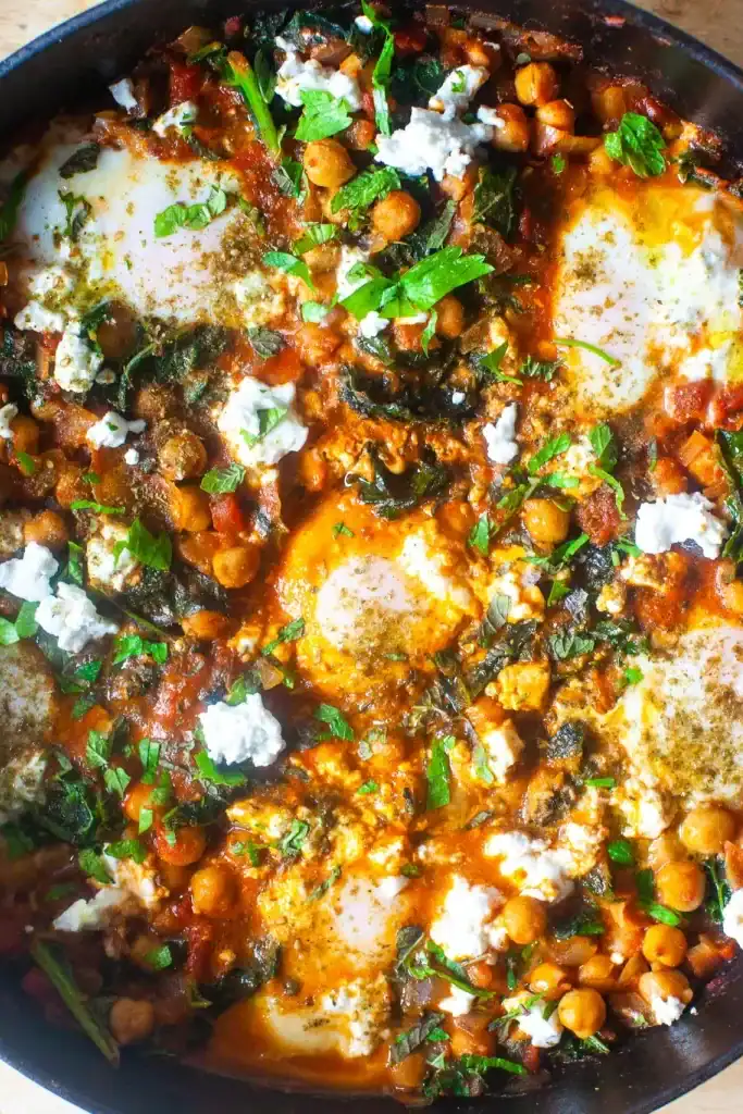 Overhead shot of shakshuka with eggs, chickpeas and kale in a pan.