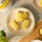 Sweet lemon bars with a buttery crust on a white plate with green trim, served with fresh herbs and lemons, perfect for homemade desserts or afternoon snacks.