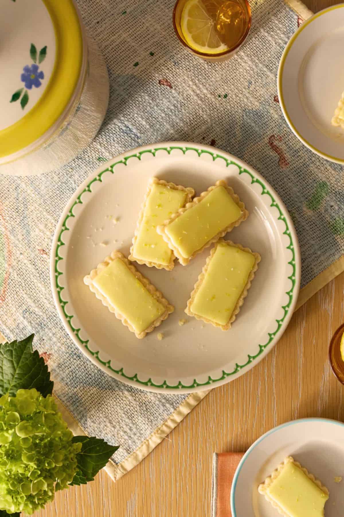 Rich lemon bars on a decorative plate, surrounded by lemon ice tea, fresh flowers, and vintage dishware, perfect for a fresh, homemade dessert experience.